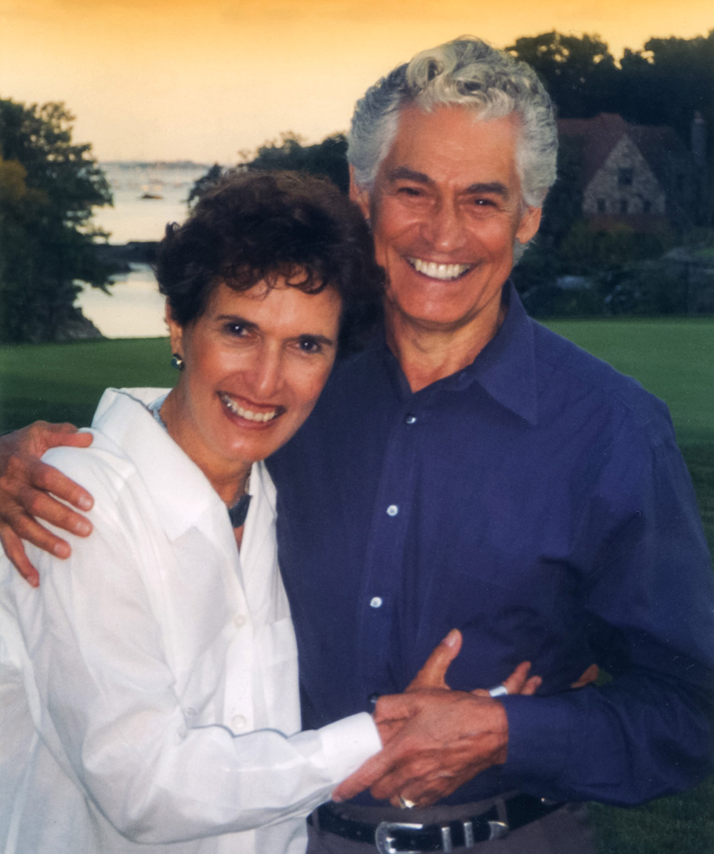 Portrait photograph of Samuel Sporn '53 (right) in a dark navy blue button-up dress shirt and Ellen Sporn (left) in a white button-up dress shirt smiling and posing for a picture together hugging each other outside in the late evening