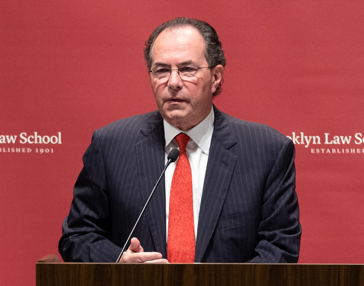 Portrait headshot photo of Frank Aquila at a podium stand that has a microphone near him as he is standing in front of a red backdrop background showing the Brooklyn Law School typography letters logo along with the established year 1901; He is wearing dark black-colored outer frame see through prescription glasses, a dark black plaid business suit blazer, white button-up dress shirt underneath, and a faded gradient red colored tie equipped