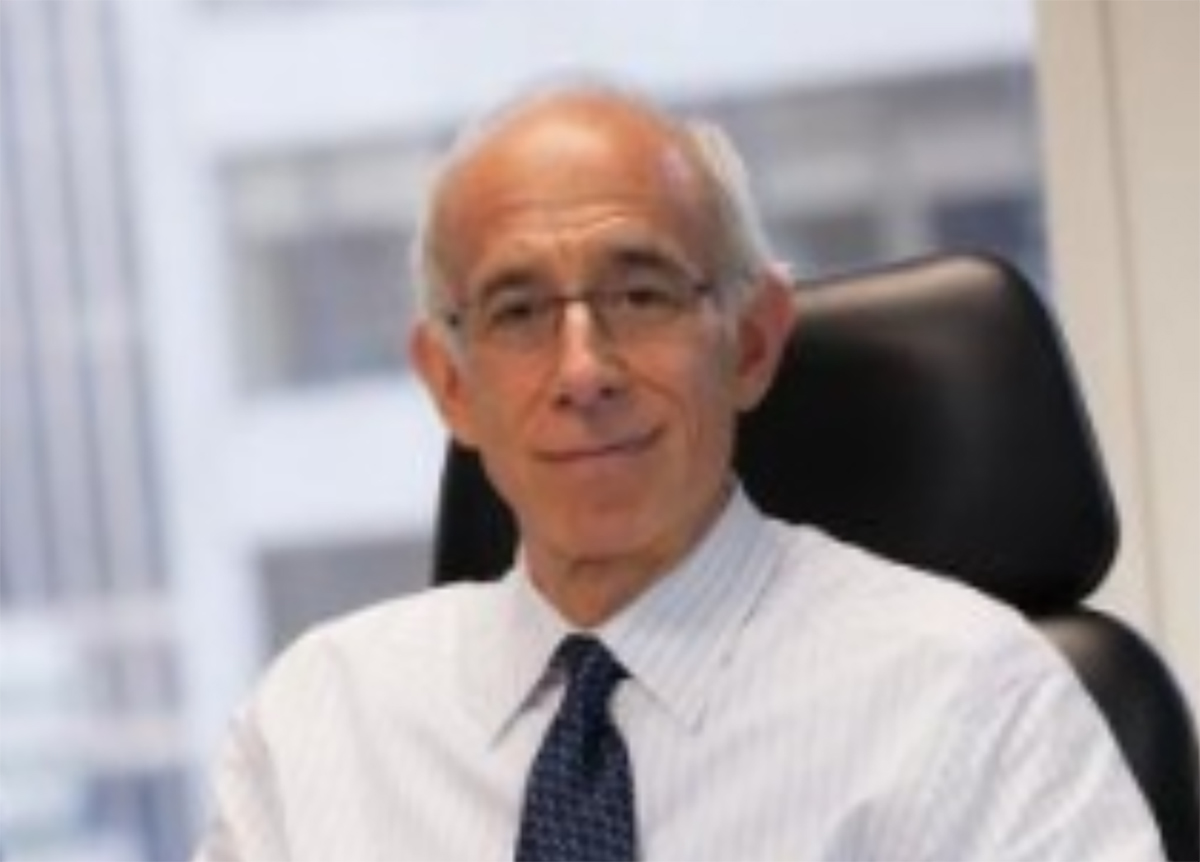 Portrait headshot photo of Noah Hanft grinning in chrome-colored tinted outer frame see through prescription glasses, white button-up plaid dress shirt, and a dark navy blue tie equipped as he is seated in a black office chair