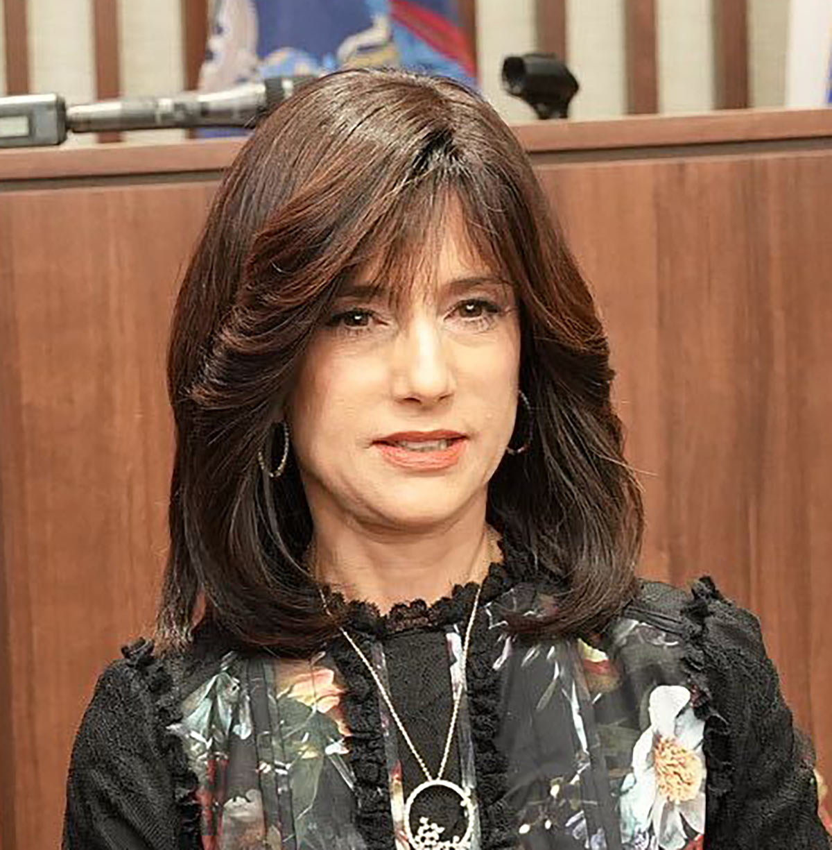 Portrait headshot photo of Hon. Rachel Freier grinning in a black blouse dress that has floral illustrative decorations on it and has on a necklace with earrings equipped as she is standing somewhere inside a courtroom setting area