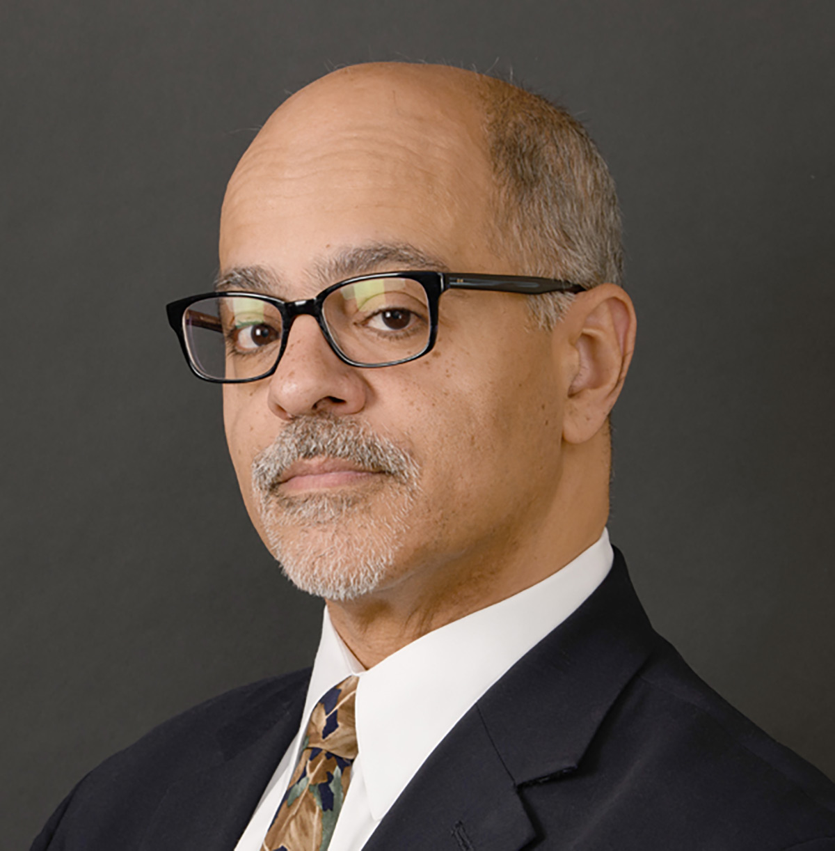 Portrait headshot photo of Hon. Ramon E. Reyes Jr. grinning in dark black-colored outer frame see through wayfarer prescription glasses that have a chrome-colored inner frame tint, a black business suit blazer, white button-up dress shirt underneath, and a multi-colored (dark blue/dark brown/dark forest green) plus leaf shape pattern tie equipped