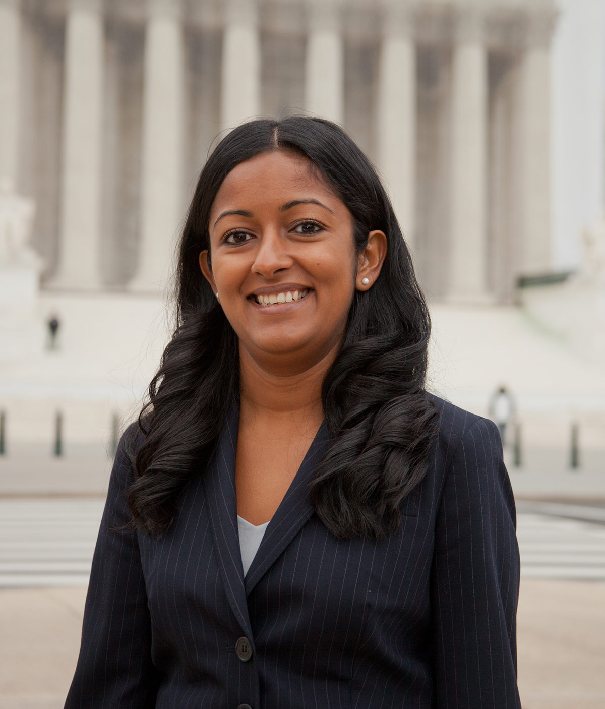 Portrait headshot photo of Sparkle L. Sooknanan smiling in a black plaid business suit blazer, a dark grey blouse/dress underneath, and has on pearl white circular shaped earrings as she is standing outside nearby the front of the United States Supreme Court building